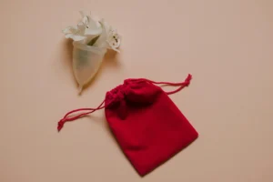 Minimalist image of a menstrual cup with flowers on a pink background, alongside a red bag.
